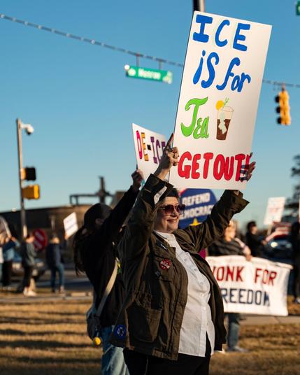 A person in sunglasses and an olive-green jacket stands in a line of protesters on a sunny street corner, holding a large handmade sign over their head that reads “ICE IS FOR Tea GET OUT!” with a drawing of a glass of iced tea, while other demonstrators and banners, including one that says “HONK FOR FREEDOM,” are visible in the background.