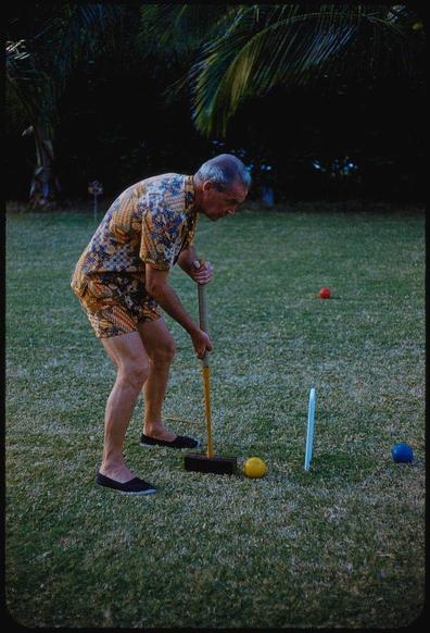 The image features an older man in a vibrant Hawaiian print shirt and shorts playing croquet on a grassy lawn. He is bending over, using his mallet to strike the ball towards two colored balls lying ahead of him. The setting appears to be outdoors during dusk or early evening as indicated by the dim lighting. Surrounding vegetation includes palm trees suggesting a tropical location. In the background, there are more colorful items like an orange and blue plastic object on the ground which could possibly belong to another game or activity.

Additional information provided mentions that this photograph is from Mill Reef Club in Antigua, Guatemala taken during January 1959 by Toni Frissell for Sports Illustrated magazine under a series titled "Picnic to Venture." This image captures one of her early works as part of an assignment. Notably, she was among the first female photographers featured regularly on Life Magazine and known primarily for sports photography.

In terms of keywords: The photograph is associated with Mill Reef Club in Guatemala; it's from November 12, 1963 when Toni Frissell contributed to Sports Illustrated magazine about an assignment involving a picnic adventure.