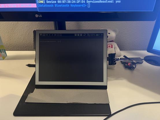 Photo of a PineNote tablet propped up on a desk below some monitors, connected via a jury-rigged UART dongle to a USB hub. The PineNote is currently displaying a terminal.