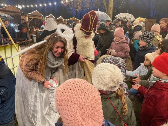 Gruppe von Menschen, darunter Kinder in Winterkleidung, steht um eine Person mit Engelsflügeln und einem weißen Gewand. Im Hintergrund ist ein festlich beleuchteter Stand zu sehen.