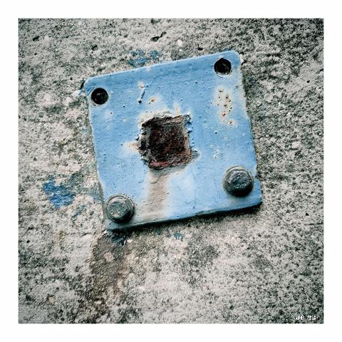 Quadratisches Farbfoto, das eine quadratische blaue Metallplatte mit vier Bohrungen zeigt, die mit zwei Schrauben am ziemlich verwitterten Betonboden angeschraubt ist.
