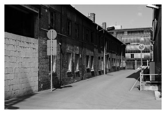 Eine leere, schmale Straße zwischen alten Backsteinfabriksgebäuden. Die Fenster der langen Hauswand links wirken verwittert, einige sind beschädigt. Am Ende der Gasse steht ein moderneres Gebäude mit großen Glasflächen und dem Schriftzug „Obermeyer“. Verkehrszeichen und Straßenlampen werfen lange Schatten in der Sonne.