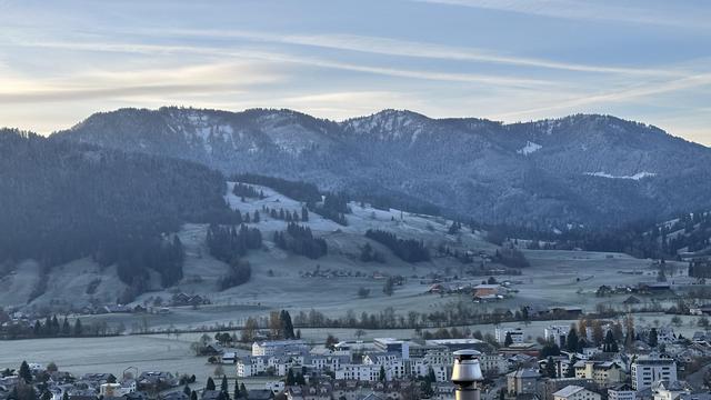 View looking south past the Swiss village of Unterägeri to the Rossberg. The valley is still mostly in early morning shadow and the higher elevations have a dusting of snow. The sky is light blue with a few clouds and contrails.