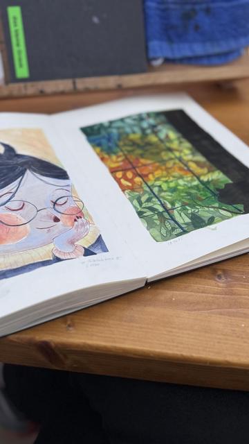 An open sketchbook on a wooden table features two pages. One page displays a whimsical illustration of a mouse character with glasses and rosy cheeks, and the other page showcases a colorful design of leaves and flowers in vibrant hues.