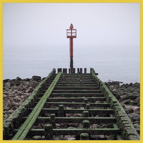 At the end of a by seaweed green colored pier stands a red pole with a harbor light. The weather is misty: you can only guess where the sea turns into air.
