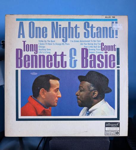A vinyl album cover standing upright against a blue wall background. The album is "A One Night Stand" by Tony Bennett & Count Basie as indicated by the larger lettering across the top of the white cover. Between the words are the tracks on the album in smaller lettering. The lower half of the cover has the two artists facing each other from the shoulders up, both rendered as a photo that's been painted over. The man on the left is white, has short dark hair and a pointed nose. He's wearing a thick red woollen jacket over a blue shirt with its collar just poking over the top. The man on the right is black, wearing a small, brimmed, black hat and just a little grey hair showing underneath. He has a trim moustache and is wearing a white shirt with striped blue tie.