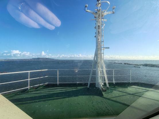 Picture from the viewing bridge of an ocean-going car ferry, looking out over a blue sea and azure sky. A sister ferry is visible in the distance. A rocky breakwater is visible on the right of frame. A small island connected by an isthmus to the mainland is visible on the left of frame.