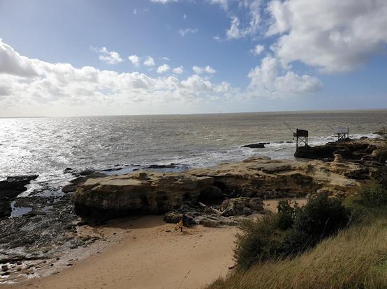 Côte rocheuse la mer et une plage au premier plan, et un carrelet au loin.