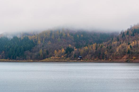 Das Bild zeigt eine neblige Herbstlandschaft an einem Gewässer, wahrscheinlich einem See oder Stausee, umgeben von bewaldeten Hügeln.
Vordergrund: Das Wasser nimmt den unteren Teil des Bildes ein. Es ist dunkel und ruhig, mit einer leicht strukturierten, grauen Oberfläche, die die melancholische Stimmung verstärkt.
Mittelgrund: Direkt am Ufer steht ein kleines, dunkles Haus oder eine Hütte. Es wirkt isoliert und fügt der ansonsten reinen Naturkulisse einen menschlichen Akzent hinzu. Die Hütte steht auf einem kleinen, flachen Streifen Land, bevor der Wald steil ansteigt.
Hintergrund: Der Hügel ist dicht bewaldet und zeigt die typischen Herbstfarben. Man sieht eine Mischung aus Nadelbäumen (dunkelgrün) und Laubbäumen, deren Blätter in leuchtenden Gelb-, Orange- und Rosttönen gefärbt sind. Die Farben bilden einen warmen Kontrast zu dem ansonsten gedämpften Licht.
Stimmung: Die Spitze des Hügels und der Himmel sind in einen dicken, weißen Nebel oder Dunst gehüllt, der die Konturen weicher macht und eine geheimnisvolle, fast mystische Atmosphäre schafft. Das Licht ist weich und diffus, was auf einen frühen Morgen oder einen bewölkten Tag hindeutet.
Zusammenfassend strahlt das Bild eine Atmosphäre der Ruhe, Abgeschiedenheit und kühlen Herbststille aus.