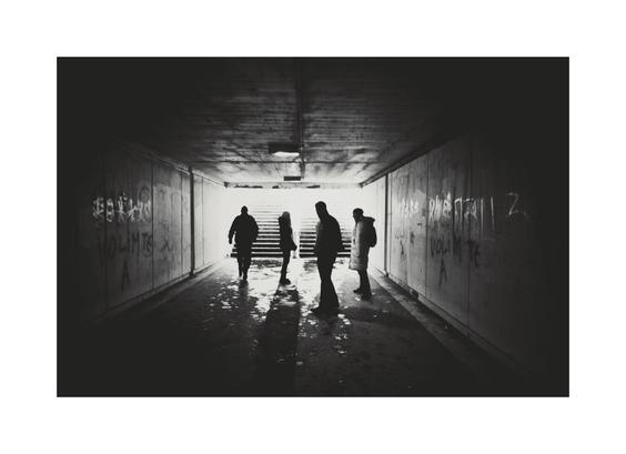A low-angle, black and white photograph of four people in silhouette walking down a concrete pedestrian tunnel toward a bright light at the end. The tunnel walls are covered in graffiti ("VOLIM TE" meaning "I love you" in Serbian can be seen written on both sides of the tunnel. The ground is wet and reflective, with puddles reflecting the figures. The figures are facing away from the camera, except for one, casting long shadows.