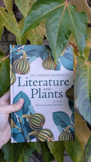 Holding the Cambridge Handbook of Literature and Plants in front of a wall overgrown with large green leaves