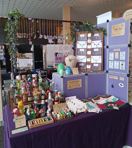 A shop stall, with a purple setup (tablecloth and shelf).
The stall is decorated with leafs.
The items that are presented are: Potionbottles, Corsets, Necklaces and Artprints.