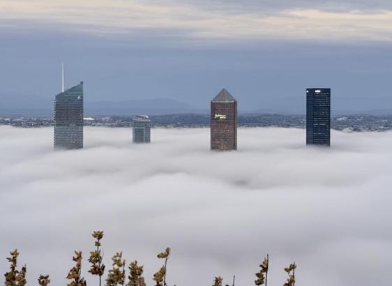 Vue de profil et probablement à partir de la cathedrale de Fourvière de l'hypercentre de la ville de Lyon par temps gris. Le brouillard est intense et couvre l'ensemble de la ville. Seules, les tours les plus hautes dépassent cette nappe brumeuse intense ce qui souligne la hauteur des édifices par rapport au reste de la cité. Effet surréaliste. 4 tours dépassent. L'une à la forme d'un crayon car son sommet est recouvert d'une pyramide en verre qui lui donne l'apparence d'une mine de crayon d'où son surnom.