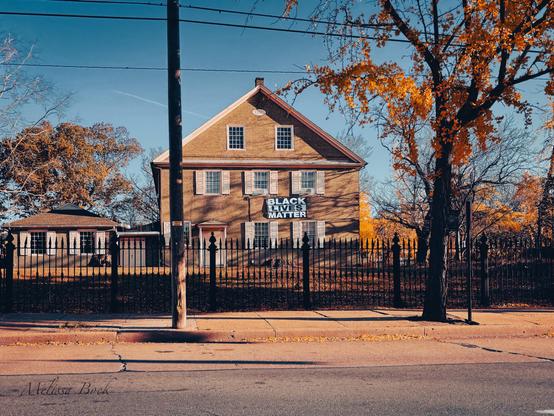 A stately brick building, set back from the road and behind a wrought iron fence. The building bears a large banner that reads “Black Lives Matter” in a block font.