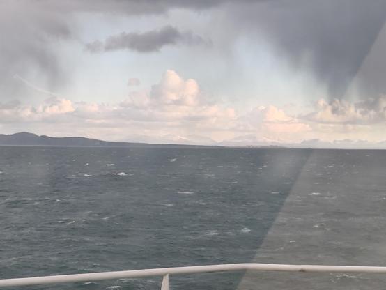 Picture taken from a ship, looking out to land, with the head of a peninsula in the foreground and snow covered mountains of North Wales in the distance. The sea is green-grey, with the occasional white crest.