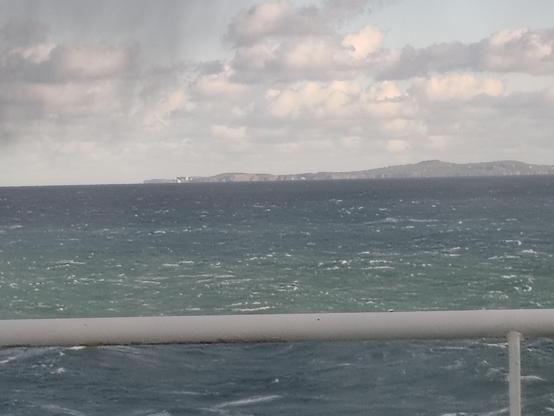 Picture of a distant lighthouse at the base of a headland. The sunlight makes the white lighthouse stand out against its background. The sea has small white crests on wave tops.