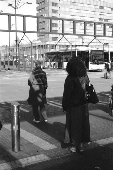 Two people waiting separately at a bus station, with their backs turned towards the viewer. The girl closest to the viewer wears a long dark coat, a shoulder bag and curly black hair. In front of her is an older lady with grey hair. The sun casts strange shadows across their backs. In the background, a bus is waiting in front of an apartment building.