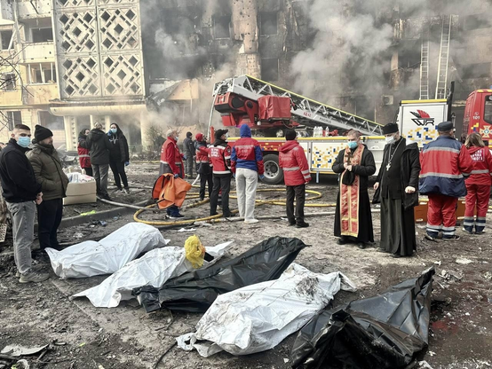 Corpses of civilians killed by ruzzian attack, covered by white sheets. Rescuers and survived victims of attack seen in front of damaged apartment building. 
There is some smoke visible.
Rescuing works are still in progress.