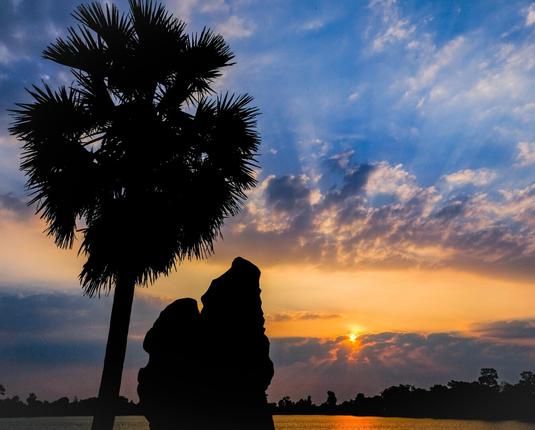 Photo shows a sunrise over a lake framed by a shoreline of forest to the right and the silhouette of a palm tree and a statue of guardian lion to the left. The rising sun is about 30 minutes off being clear of the horizon but has risen through a layer of morning fog and clouds before reaching a height with deep blue sky.  A bit higher on this morning sky is another belt of clouds. In the field between the son face is sending rays up and down in the cloud systems, on a background of red and yellow slowly turning into blue. The lake under the sun is coloured deep red mirroring the sun, changing into liquid golden colour off the direct sun trail. Cambodia is as safe, comfortable and kind as ever. Angkor and Siem Reap welcomes visitors.