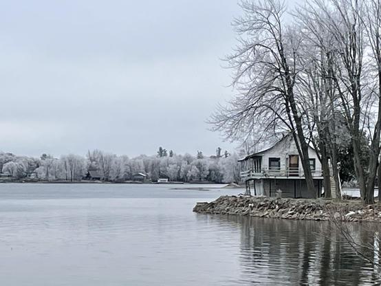 An old boathouse on the shore of a lake. The trees around it and across the lake are covered in a hard frost.