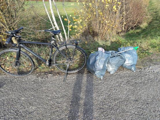 Mein schwarzes Fahrrad, rechts davon drei Säcke mit eingesammelten Müll. Die Säcke sind blau und fassen 60l, darauf liegen meine zerzausten Arbeitshandschuhe und mein Müllgreifer. Vorne sieht man etwas Terr, dahinter Grünfläche mit Bäumen, Büschen und Abhang. Ach ja, durch hecken krauchen macht auch nur bedingt Spaß, die sind hier auch noch zu sehen.