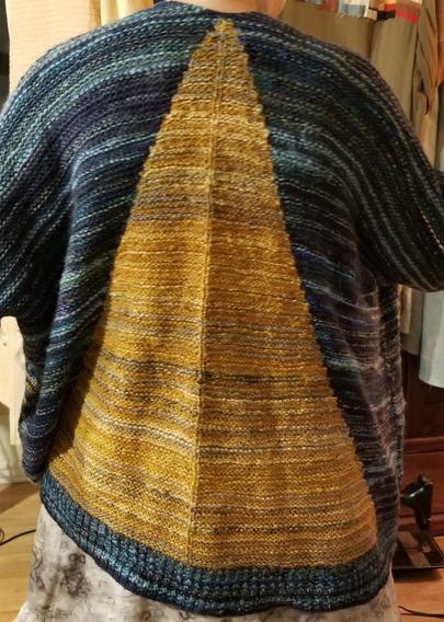 Modeled shot of the back of an oversize long sweater with a giant yellow triangle on a blue field. Because of the gauge difference between the yarns, the sweater looks very saggy.
