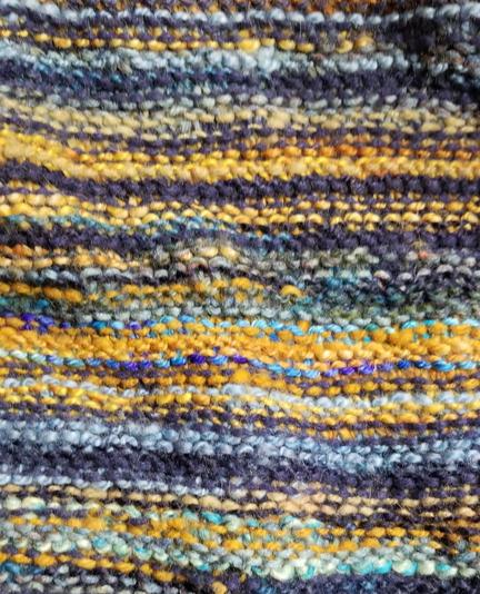 A close up of a section of knitting in garter stitch, in narrow stripes of various handspun yarns in shades of blue, gold, and orange.