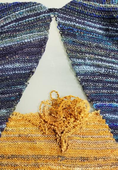 Close up of a section of knitting in garter stitch. A yellow intarsia triangle is being unraveled from within a blue background and there's a pile forming of kinky yarns. A glass tabletop is visible in the space where the knitting has been removed.