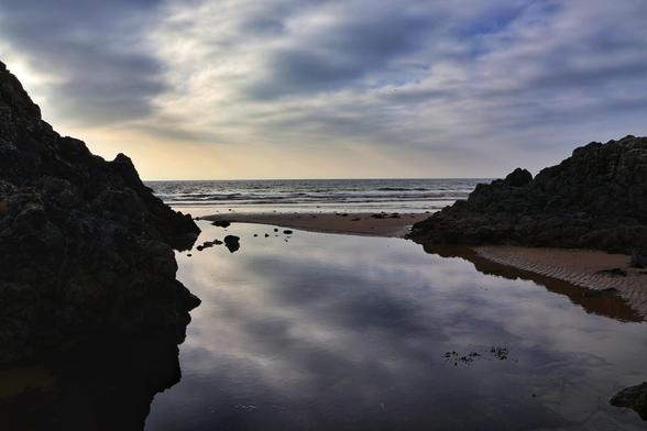 A serene coastal scene captured at what appears to be either dawn or dusk. The image showcases a tranquil, shallow tidal pool reflecting the soft hues of the sky, framed by rugged, dark rock formations on either side. Beyond the pool, a sandy beach stretches out towards the calm sea, where gentle waves lap the shoreline. The sky above is adorned with wispy, layered clouds, adding depth and texture to the scene.