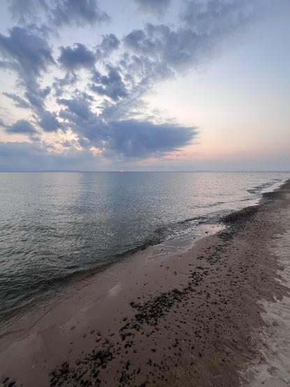 A serene beach scene at sunrise at the Whitefish Point Lighthouse on Lake Superior, featuring calm waters with gentle waves lapping at the shore. The sky is filled with soft clouds in shades of blue and pink, and a faint sunset is visible on the horizon. The sandy beach includes patches of small stones.