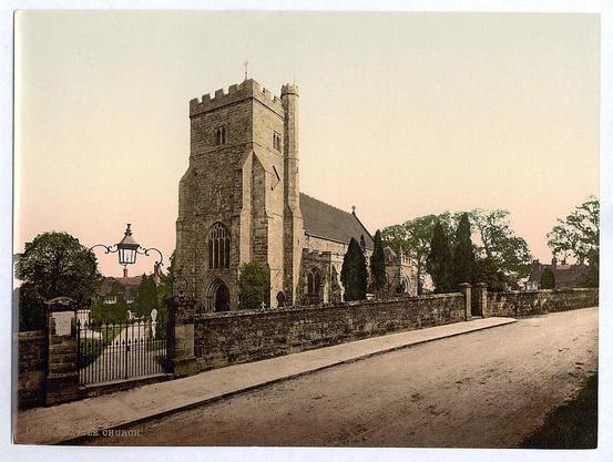 This image features a historic stone church with Gothic architectural elements, including pointed arch windows and buttresses. A tall tower rises prominently above the rest of the structure, capped by what appears to be a cross or similar religious symbol. The church is surrounded by mature trees and has an iron fence enclosing its grounds. In front of the church lies a paved path that leads up to it from a nearby road where an ornate street lamp stands on the left side of the image. A stone wall separates this pathway from what appears to be a cemetery, as indicated by headstones visible in the background. The photograph is sepia-toned and has some handwritten inscriptions, including "St. Mary's Church" at the bottom right corner, indicating its location or name.