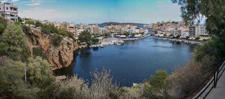 A scenic view of a harbor surrounded by buildings and rocky cliffs, with calm water reflecting the sky. There are boats moored along the shoreline, and lush greenery in the foreground.
