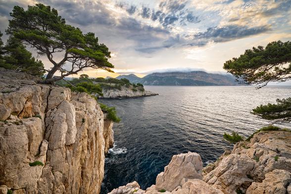 Zu sehen ist eine Meeresküste mit felsigen Steinklippen, während im Hinterrgund bei bewölktem Himmel die Sonne aufgeht.