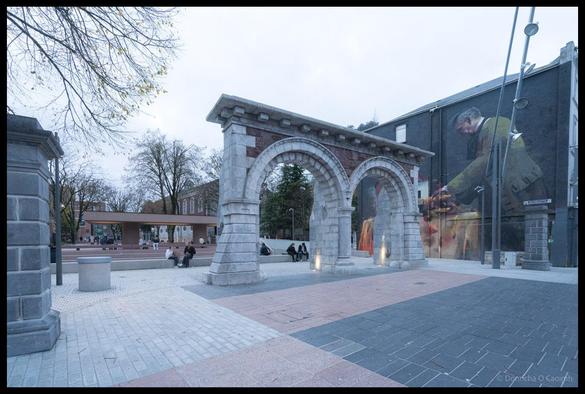 Wide-angle view of newly redesigned Bishop Lucey Park entrance in Cork showing preserved historic stone archway with ornate brick detailing, modern paved plaza with people sitting on benches, covered walkway structure in background, bare winter trees, and large photographic murals on adjacent building walls under overcast sky.