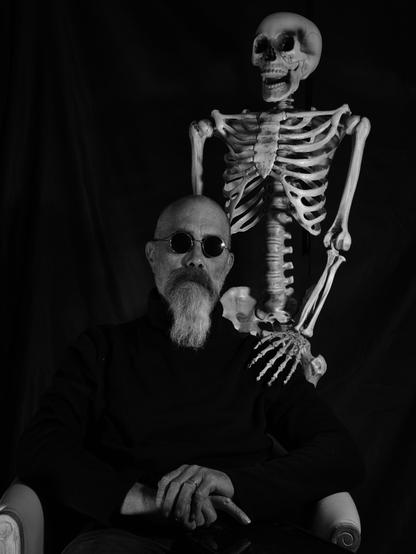 Homme avec des lunettes noires assis. derriÚre lui un squelette debout lui met la main sur l'épaule.