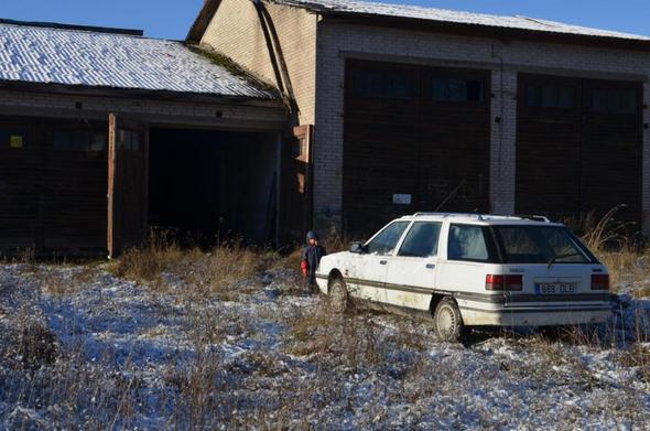 Une R21 break dans l'herbe enneigée, garée devant de vieux garages aux murs de brique blanche et toiture fibrociment. On distingue un jeune humain en combinaison d'hiver debout devant la voiture.