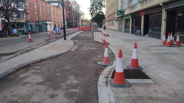 Cycleway under construction. Road left with a kerbed buffer and footway right. Cycle track does a little wiggle right then left to create a crossing refuge between the cycle track and road.