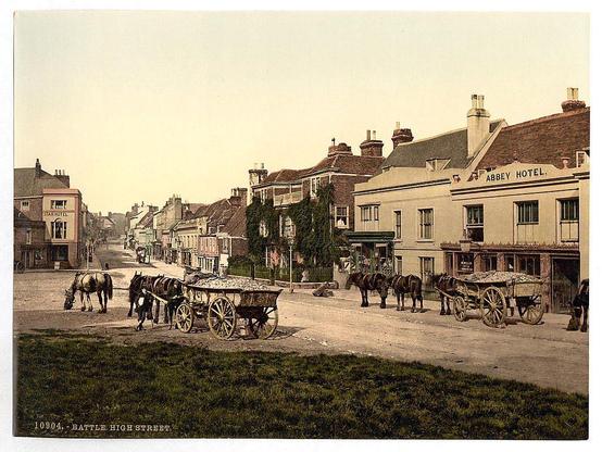 The image depicts a street scene in what appears to be an English town during the late 19th or early 20th century. The main focus is on High Street, specifically Battle High Street as indicated by the text at the bottom of the photo. The buildings lining the street are traditional British architecture with pitched roofs and various styles ranging from Victorian to Edwardian.

The foreground shows horses pulling wagons loaded with goods such as grains or hay, suggesting a market day or delivery time in an era before motor vehicles were common. The middle ground features more horse-drawn carriages moving along the road. Pedestrians can be seen walking on the sidewalks and crossing the street.

In the background, several two-story buildings display signs for hotels like "Star Hotel" and "Abbey Hotel," indicating places of accommodation for travelers or tourists. There is also a variety of shops with visible signage such as "Butchers," suggesting a mix of commercial activity along this main thoroughfare.

The overall atmosphere conveyed by the image is that of a peaceful, everyday life in a small town during an earlier time period, showcasing traditional modes of transportation and commerce typical for that era.