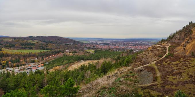Dieses Panorama zeigt eine ausgedehnte, hügelige Landschaft unter einem bewölkten Himmel, wahrscheinlich an einem kühlen Tag im Spätherbst.
Vordergrund (Rechts): Der Blick geht über einen steil abfallenden Hang, der teilweise mit trockenem Gras, niedriger Vegetation und felsigem Schutt bedeckt ist. Ein klar erkennbarer, gewundener Pfad führt den Hang hinauf und deutet auf eine Wander- oder Bergbauaktivität hin. Links davon befindet sich ein dichterer Bewuchs aus Nadelbäumen (Tannen/Fichten) und einigen Laubbäumen.
Mittelgrund: Eine grüne, bewaldete Fläche erstreckt sich über die Hügel. Eingebettet in diese Landschaft ist ein kleinerer Siedlungsteil (links), bestehend aus mehreren Gebäuden mit dunklen Dächern. Ein großes Stadtgebiet breitet sich in der Ferne aus und dominiert das Zentrum des Bildes. Man erkennt viele dicht bebaute Häuser mit orange-roten Dächern, die auf eine mittlere bis große Stadt hinweisen.
Hintergrund: Die Stadt grenzt an eine weitere hügelige Landschaft mit Ackerland und Feldern. Der Horizont wird von einem flachen, wolkenverhangenen Himmel abgeschlossen, der das Licht dämpft und der Szene eine eher ernste oder ruhige Atmosphäre verleiht.
Es ist eine Aussicht, die typisch für eine deutsche Mittelgebirgsregion ist, möglicherweise eine alte Bergbau- oder Wanderregion, von einem erhöhten Aussichtspunkt aus aufgenommen.