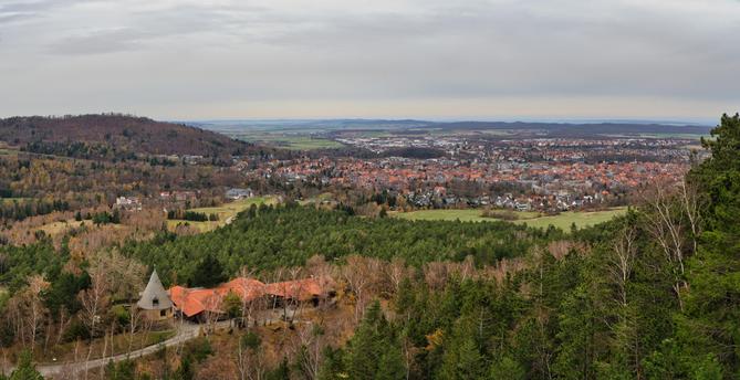 Dieses Foto zeigt eine weite Aussicht von einem bewaldeten Hügel auf eine darunter liegende Stadt, ähnlich der vorherigen Aufnahme, aber aus einem leicht anderen Winkel und mit einem markanten Gebäude im Vordergrund.
Vordergrund: Der Blick geht über einen lichten Wald, hauptsächlich bestehend aus Nadelbäumen und einigen kahlen Laubbäumen (Birken). Ein einzigartiges Gebäude sticht hervor: Es hat ein großes, geschwungenes Dach mit orange-roten Ziegeln, das sich in die Landschaft einschmiegt, und einen auffälligen, turmartigen Anbau mit einem spitzen, kegelförmigen Dach. Eine Straße oder ein Weg führt auf dieses Gebäude zu.
Mittelgrund: Eine weite Ebene trennt den Aussichtspunkt von der eigentlichen Stadt. Hier liegen Wiesen, Felder und Wäldchen. Im linken Bildteil erhebt sich ein dicht bewaldeter Hügel.
Hintergrund: Die Szene wird von einer ausgedehnten Stadt dominiert, deren Zentrum durch eine hohe Dichte von Gebäuden mit den typischen orange-roten Dächern charakterisiert wird. Dahinter erstreckt sich flaches Ackerland bis zum Horizont.
Atmosphäre: Der Himmel ist dicht bewölkt und grau, was der Szene eine ruhige, herbstliche oder frühwinterliche Stimmung verleiht. Die Farben sind gedämpft, aber die Rottöne der Dächer und das Grün der Nadelbäume setzen Akzente.
Das markante Gebäude im Vordergrund legt nahe, dass es sich um ein Ausflugslokal, ein Hotel oder ein historisches Bauwerk in einem Erholungsgebiet handeln könnte, das als Aussichtspunkt dient.