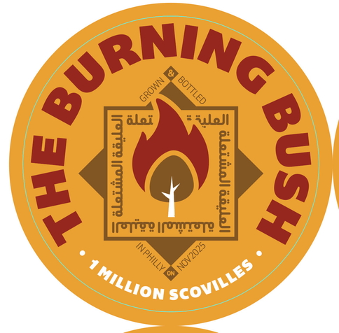 A circular orange label for "The Burning Bush" with bold red text arcing the top. Central design includes a red flame icon containing a white tree-like symbol, surrounded by Arabic script within a brown border. Above the flame: "GROWN & BOTTLED"; below: "IN PHILLY ON NOV 2025"; bottom arc: "1 MILLION SCOVILLES" in white text.