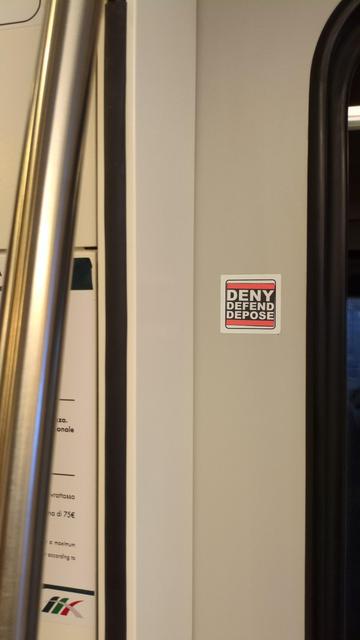 Adesivo quadrato dedicato a Luigi Mangione su treno: scritta bianca "DENY DEFEND DEPOSE" su sfondo nero. Sopra e sotto la scritta ci sono due bande rosse a ricordare il design tipico degli adesivi FCK NZS.