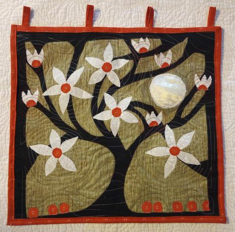 A quilted wall hanging about 24" square with a red border, black silhouette of a tree, white flower petals and red flower centers, and some red buds on the ground around the tree. The background is a brownish-gold. The hanging tabs are red. A full moon rises between the tree branches on the right.