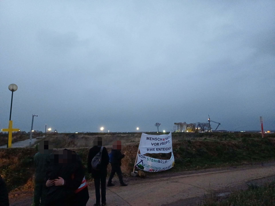 Ein paar Demonstrant*innen versammeln sich vor dem Erdwall am Werksgelände des Tagebau Hambach. Auf einem Banner steht: "Mensch & Natur vor Profit - RWE enteignen!"; im Hintergrund die Räumung des "Sündenwäldchens" mit Kränen, sowie ein Braunkohlebagger im Abendlicht.