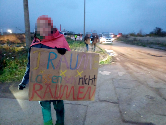 Eine Demonstrantin trägt ein Pappschild mit bunter Schrift: "Träume lassen sichnicht räumen", im Hintergrund weitere Demonstrant*innen und Polizeifahrzeuge