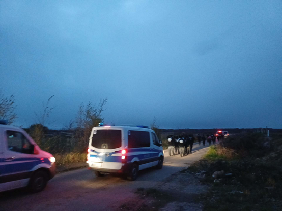 Polizist*innen zu Fuß und mit Transportern begleiten eine Demonstration auf einer dunkelen Landstraße