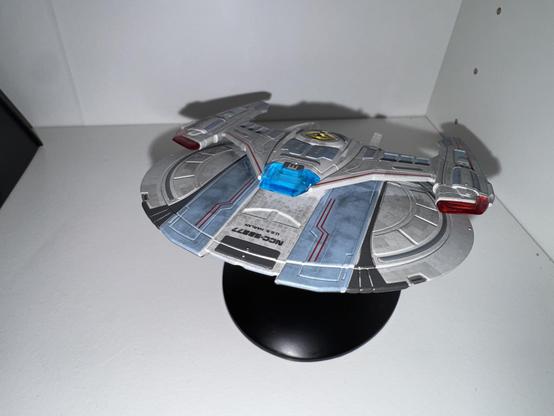 A detailed model of a futuristic spaceship, featuring a silver and blue design with red lights, displayed on a black stand against a plain background.