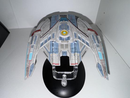 A detailed model of a starship is displayed on a black stand. The ship features a sleek, multi-colored design with blue, gray, and red accents, and includes intricate panel details. The backdrop is plain, highlighting the model's design.