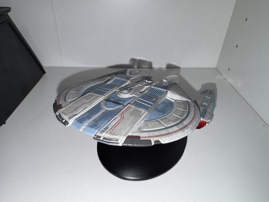 A model of a spaceship displayed on a black stand. The ship features a metallic finish with blue and silver details. It is positioned on a white shelf with a dark object partially visible in the background.