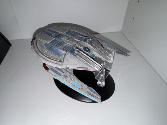 A model of a futuristic spaceship displayed on a black stand, featuring intricate detailing and a sleek design with blue, gray, and red accents. The background is a plain white surface.